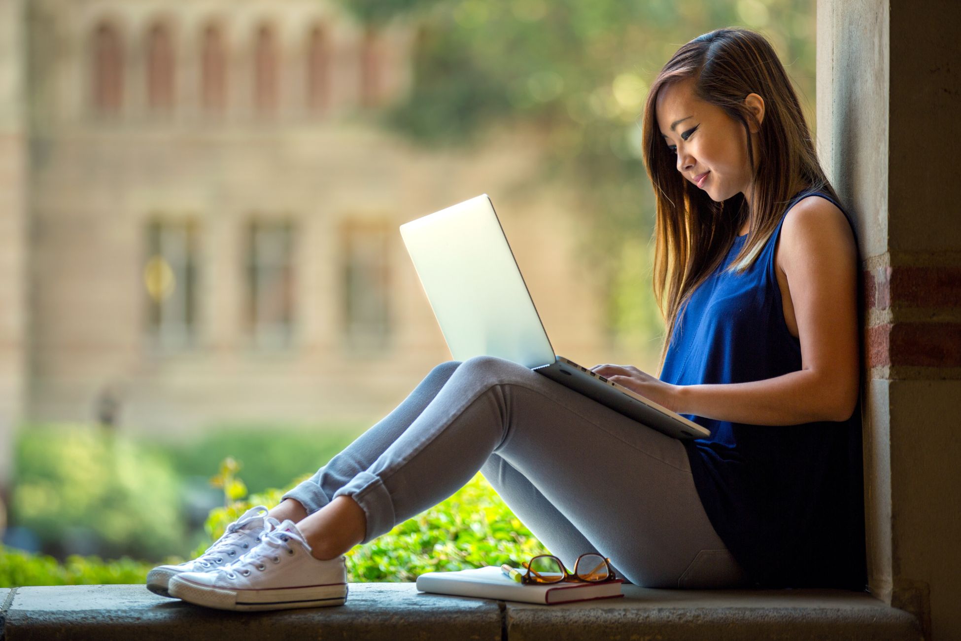 Young woman types on laptop and sits in arched wall opening with brick building in background