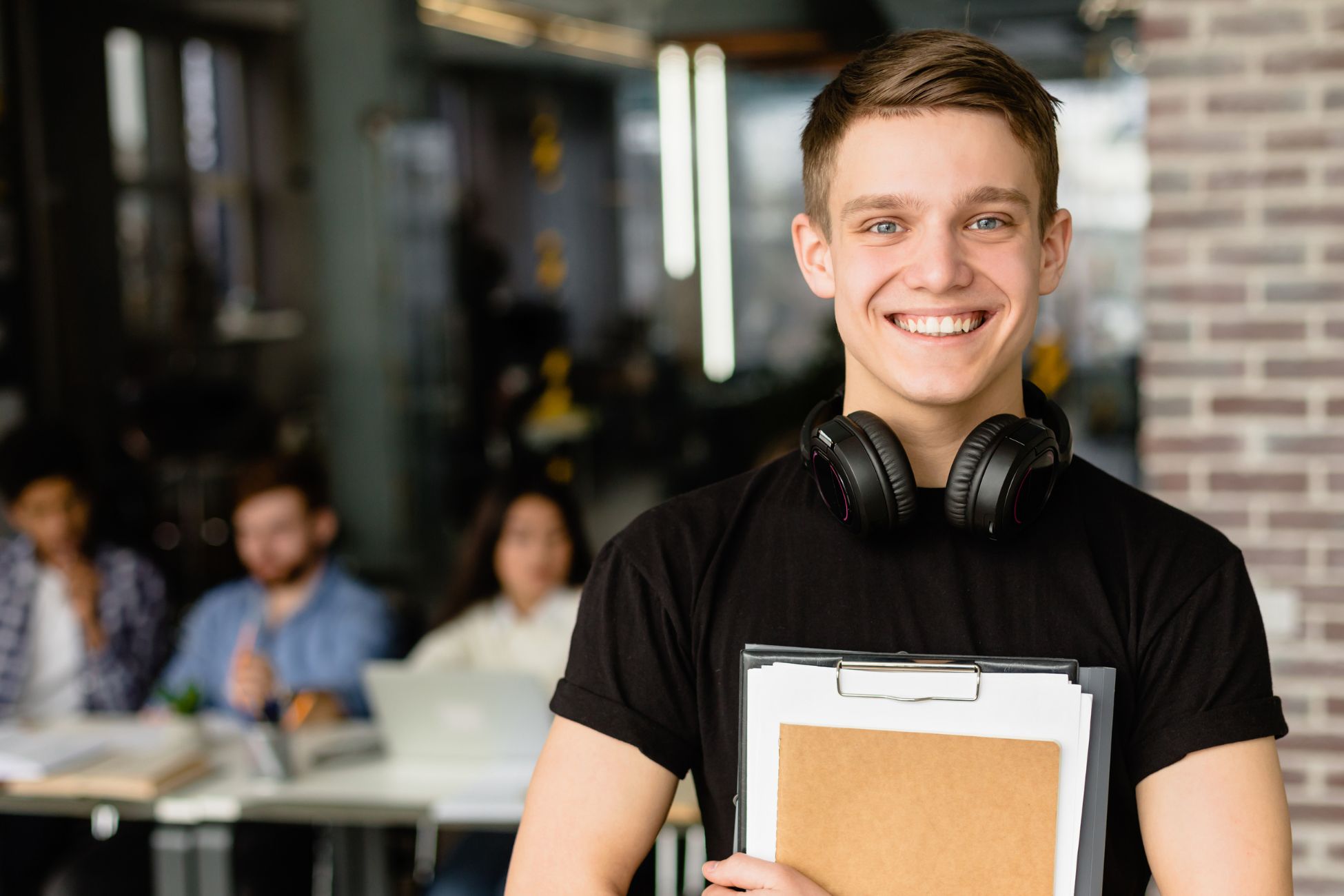 Young man with short hair stands holding a clipboard and smiles to camera; others at table behind