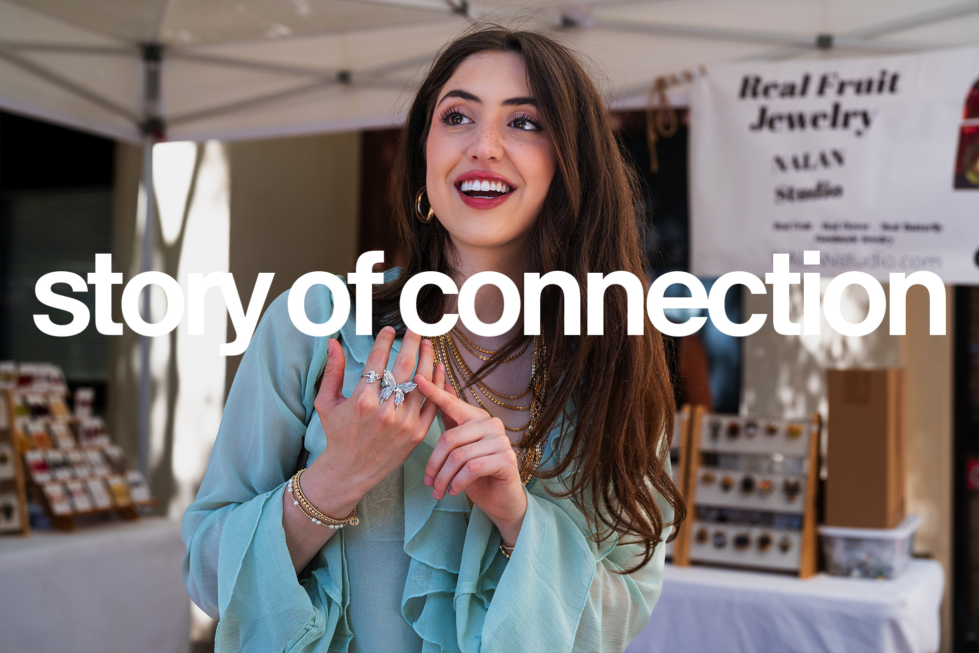 A young woman wearing gold jewelry and a light blue blouse smiles while standing at an outdoor market booth. Large white text across the image reads “story of connection.”