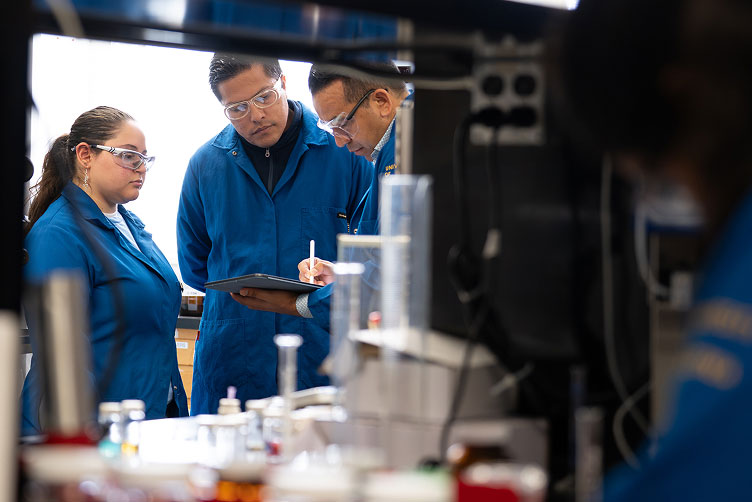 Professor Osvaldo Gutierrez holding pen and pad, with two others who are also in blue lab coats.