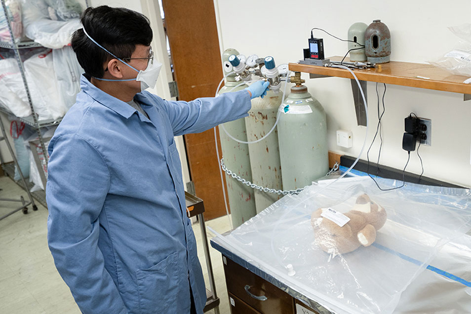 A dark haired man wearing a mask and blue lab coat measures smoke damages from a stuff toy